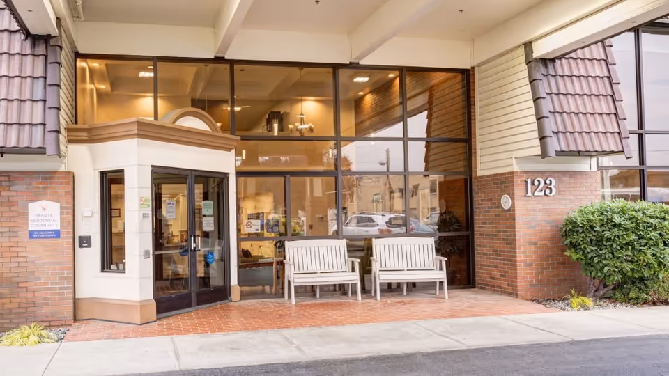 Entrance to a residential community building with large glass windows, double glass doors, two white benches on a brick patio, and the number 123 displayed on the brick wall. There is a small bush to the right and a sign on the left indicating it is a private residential community.