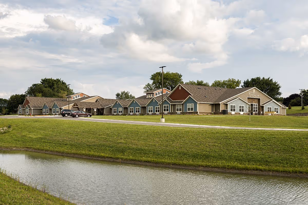 Long single-story senior living facility with multiple gabled sections, a driveway and parked cars, a grassy lawn and a pond in the foreground under a cloudy sky.
