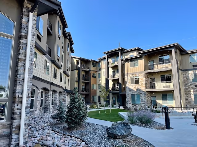 Exterior view of a multi-story senior living facility building with stone and beige siding, balconies, landscaped garden with small trees, shrubs, rocks, and a clear blue sky.