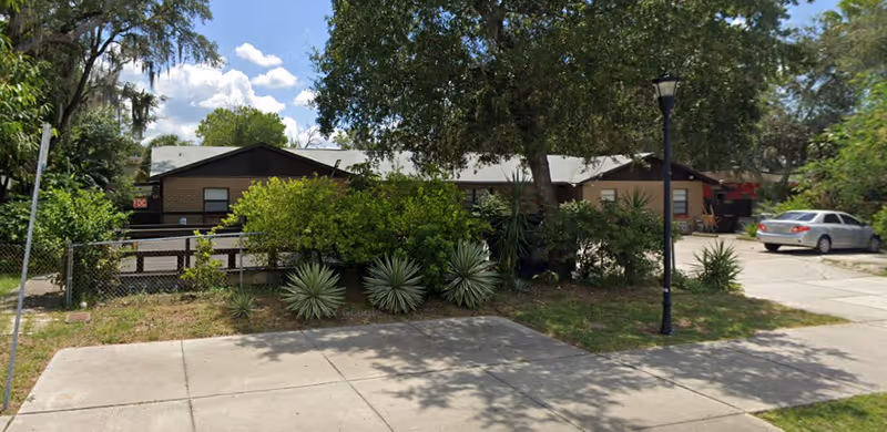 Single-story brick senior facility with trees, shrubs, a driveway, and a parked car in front.