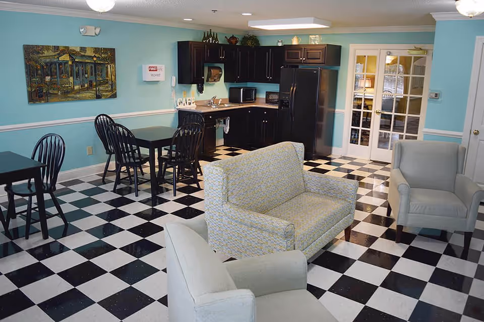 A cozy common area with black and white checkered flooring, light blue walls, and white trim. The room features a small kitchenette with black cabinets, a refrigerator, microwave, and sink. There are two small black tables with black chairs, a patterned loveseat, and two light gray armchairs. A painting hangs on the wall, and double glass doors lead to another room.