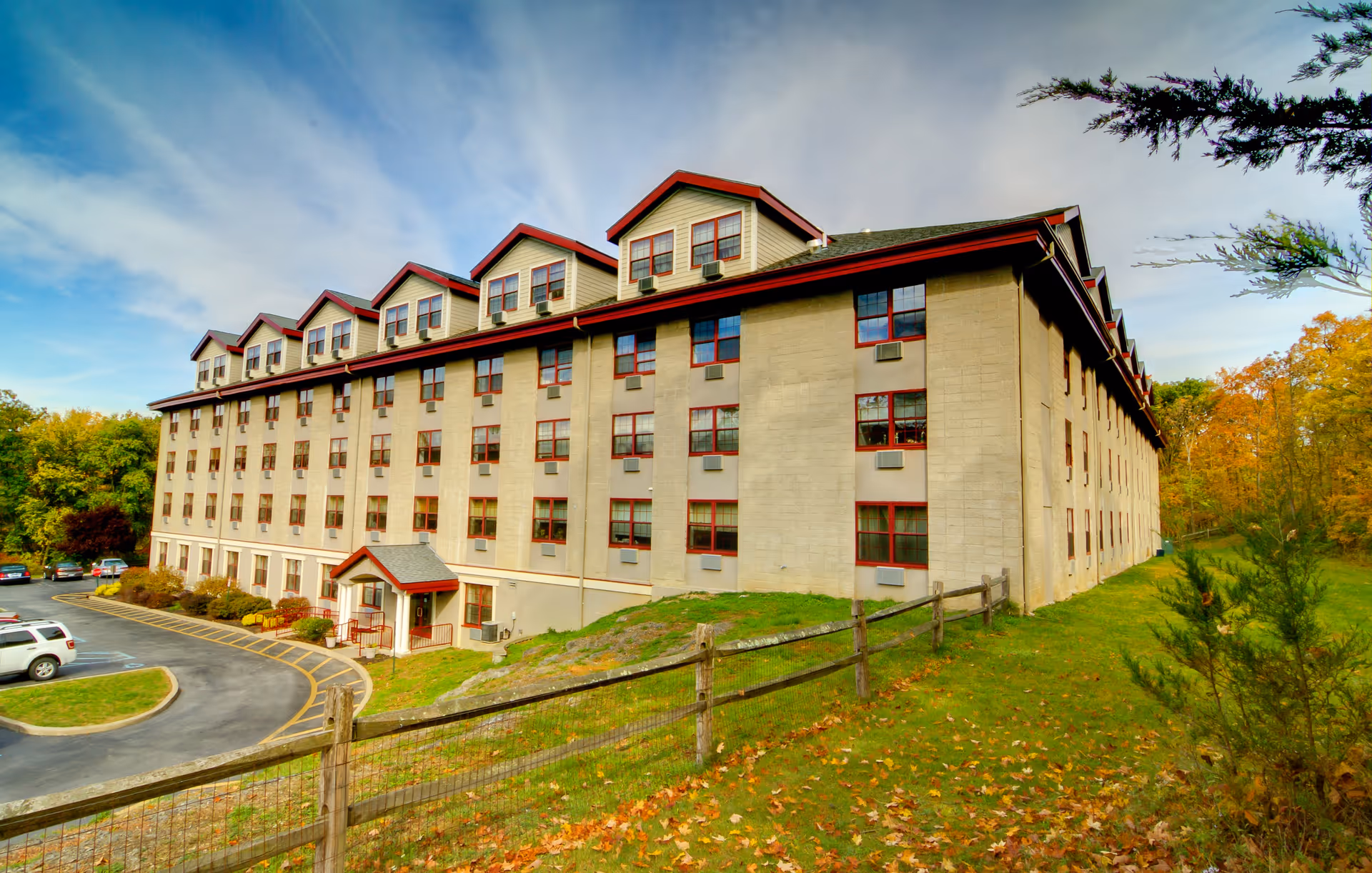 Side view of a beige multi-story senior living building with red-trim windows and autumn trees.