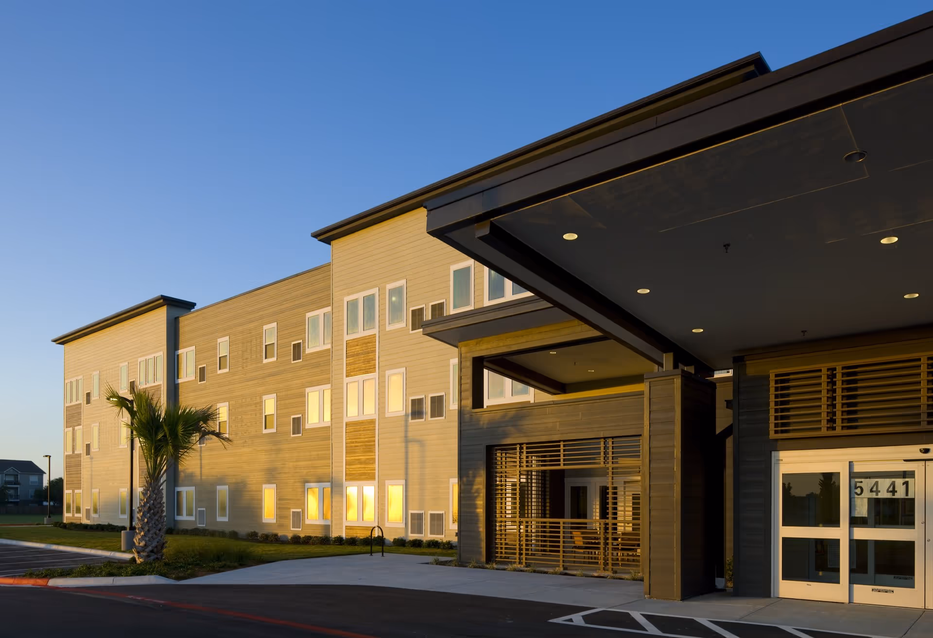 Exterior view of a modern three-story senior living facility building with multiple windows reflecting the sunset, a covered entrance with seating visible inside, a palm tree near the sidewalk, and a clear blue sky.