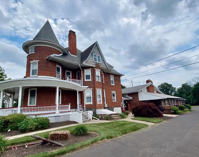 Red brick Victorian-style senior living facility with curved white porch, turret, and green lawn.