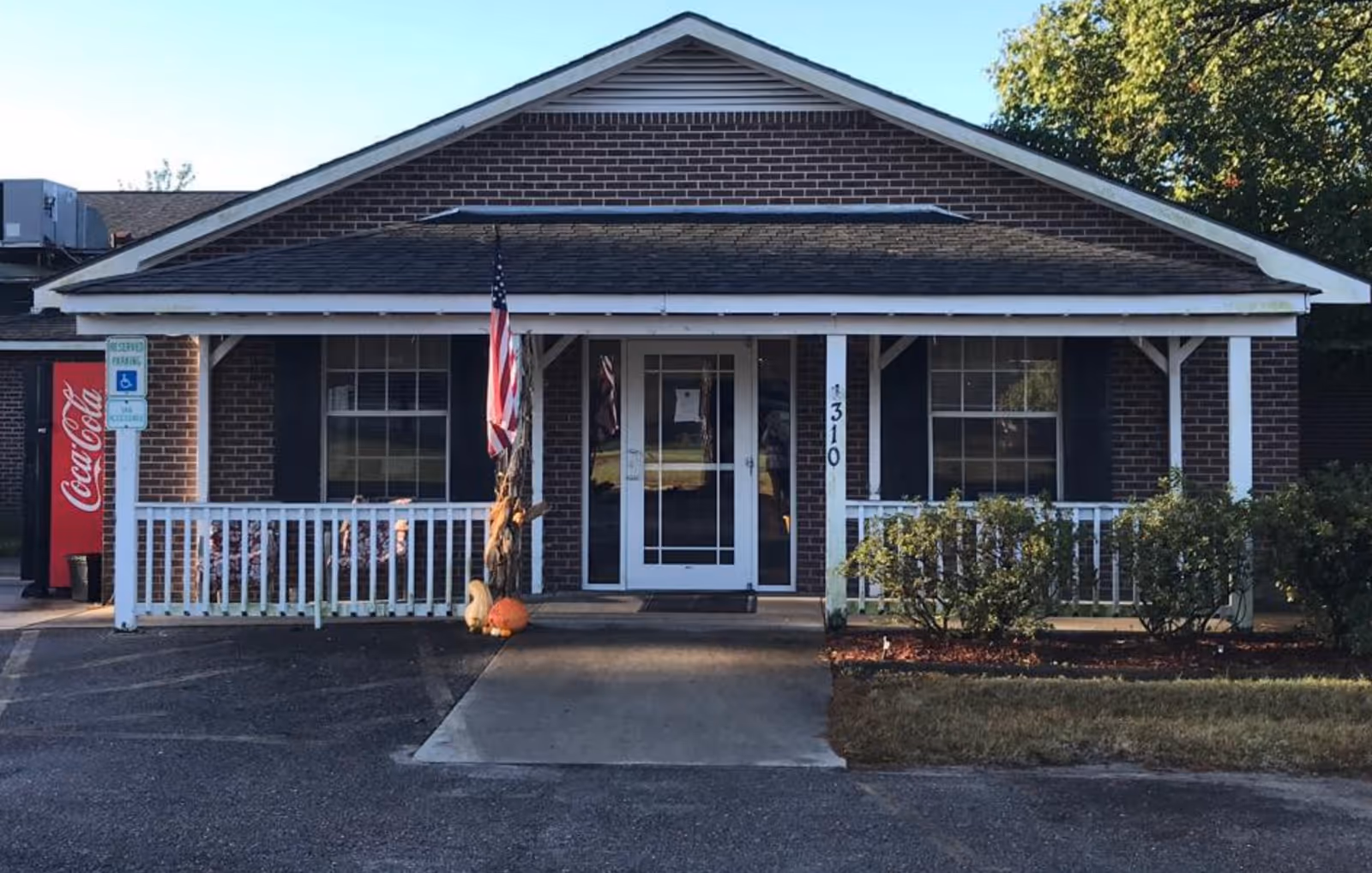 Front exterior of a single-story brick building with a covered porch, white railing, American flag, and a Coca-Cola vending machine at the left.