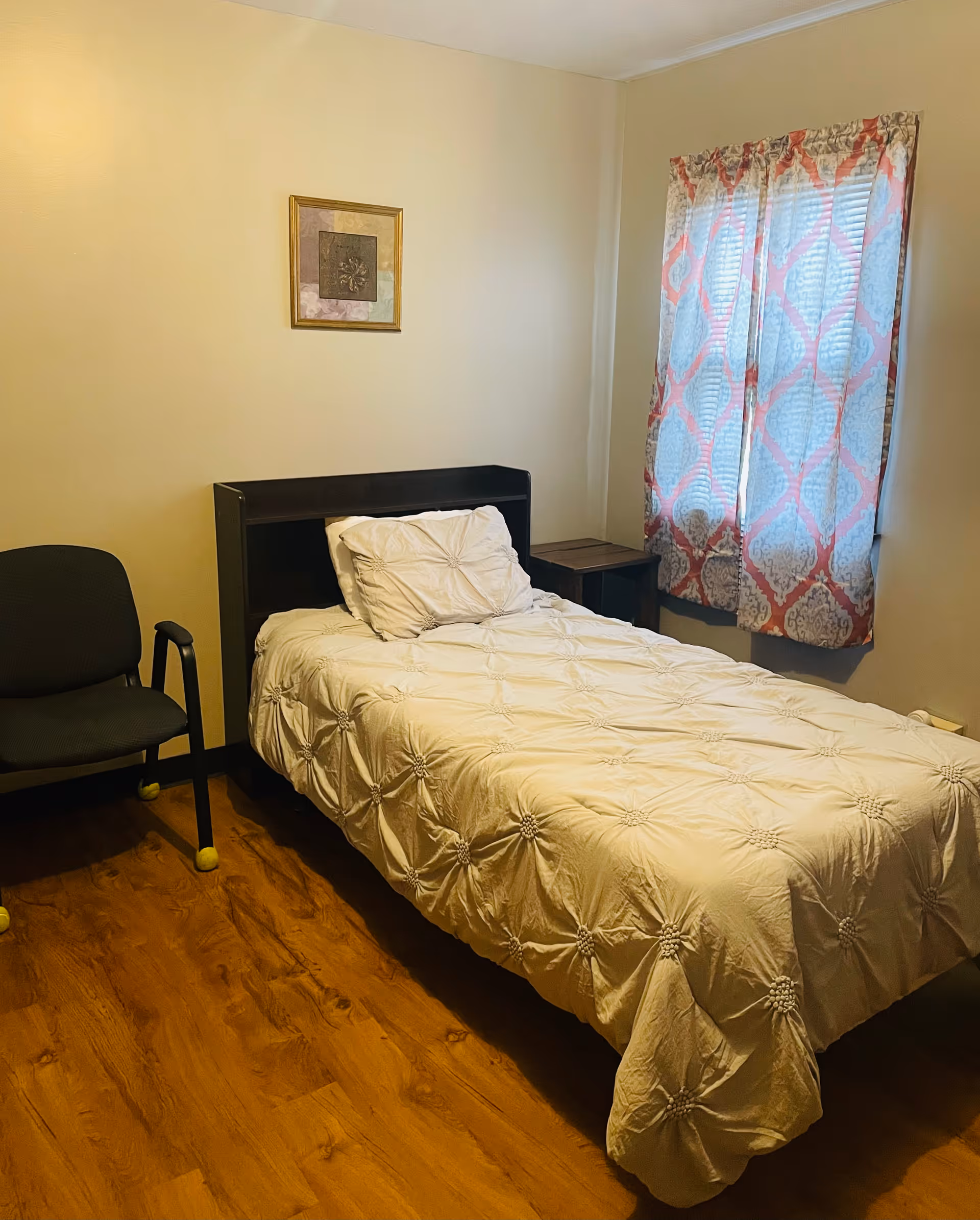 Modest bedroom with a bed, chair, nightstand, and patterned curtains.