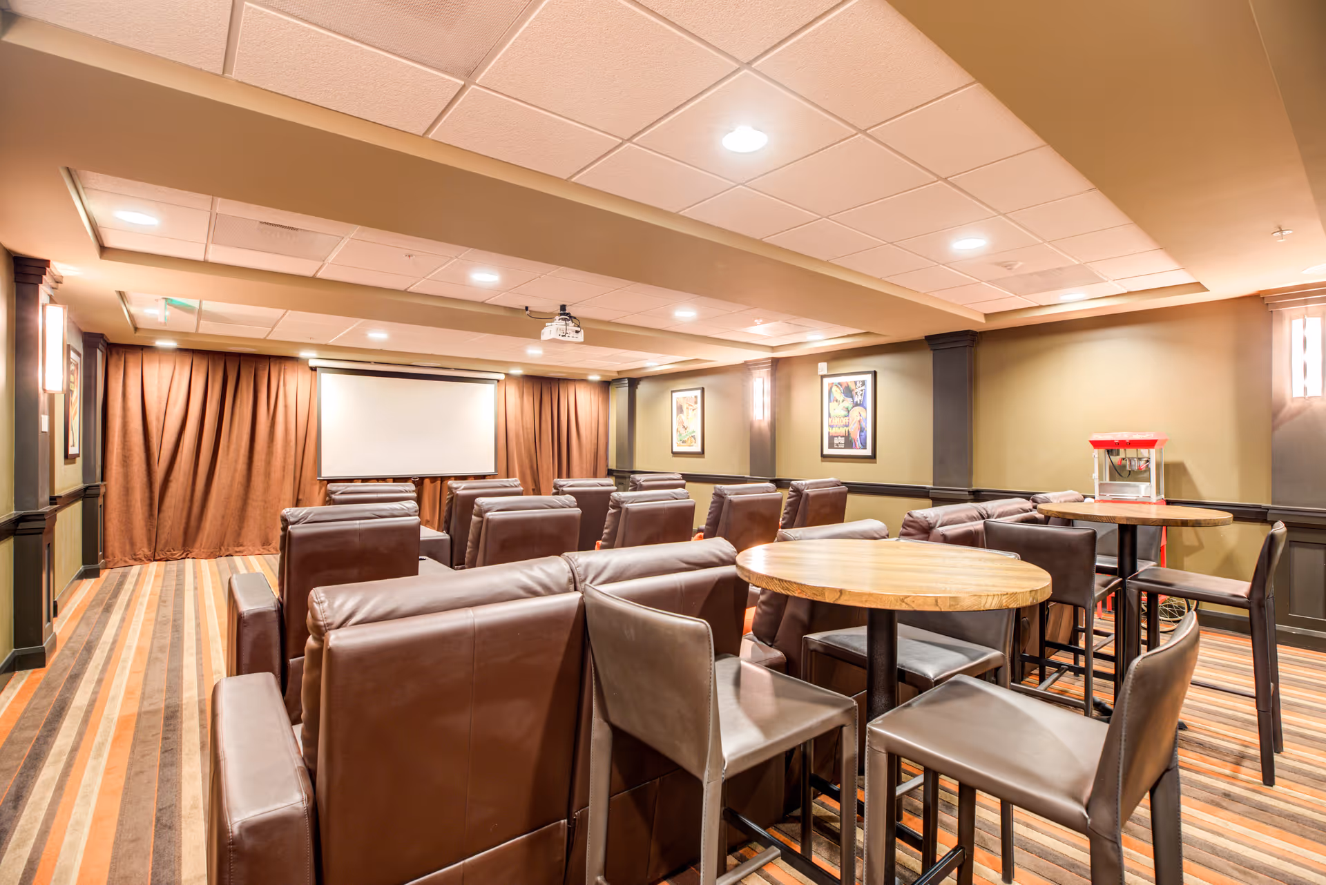 A cozy theater-style community room with rows of leather recliners facing a projection screen, high-top round tables and a popcorn machine.