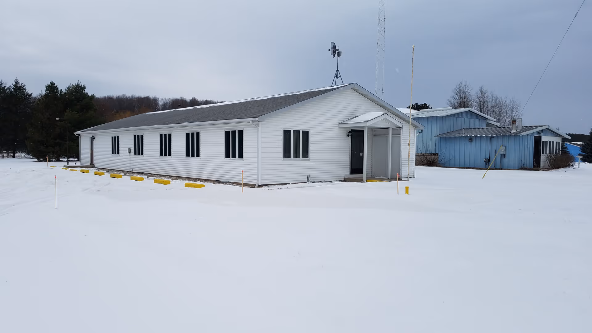 Snow-covered, single-story white facility with a small entryway and a blue building nearby.