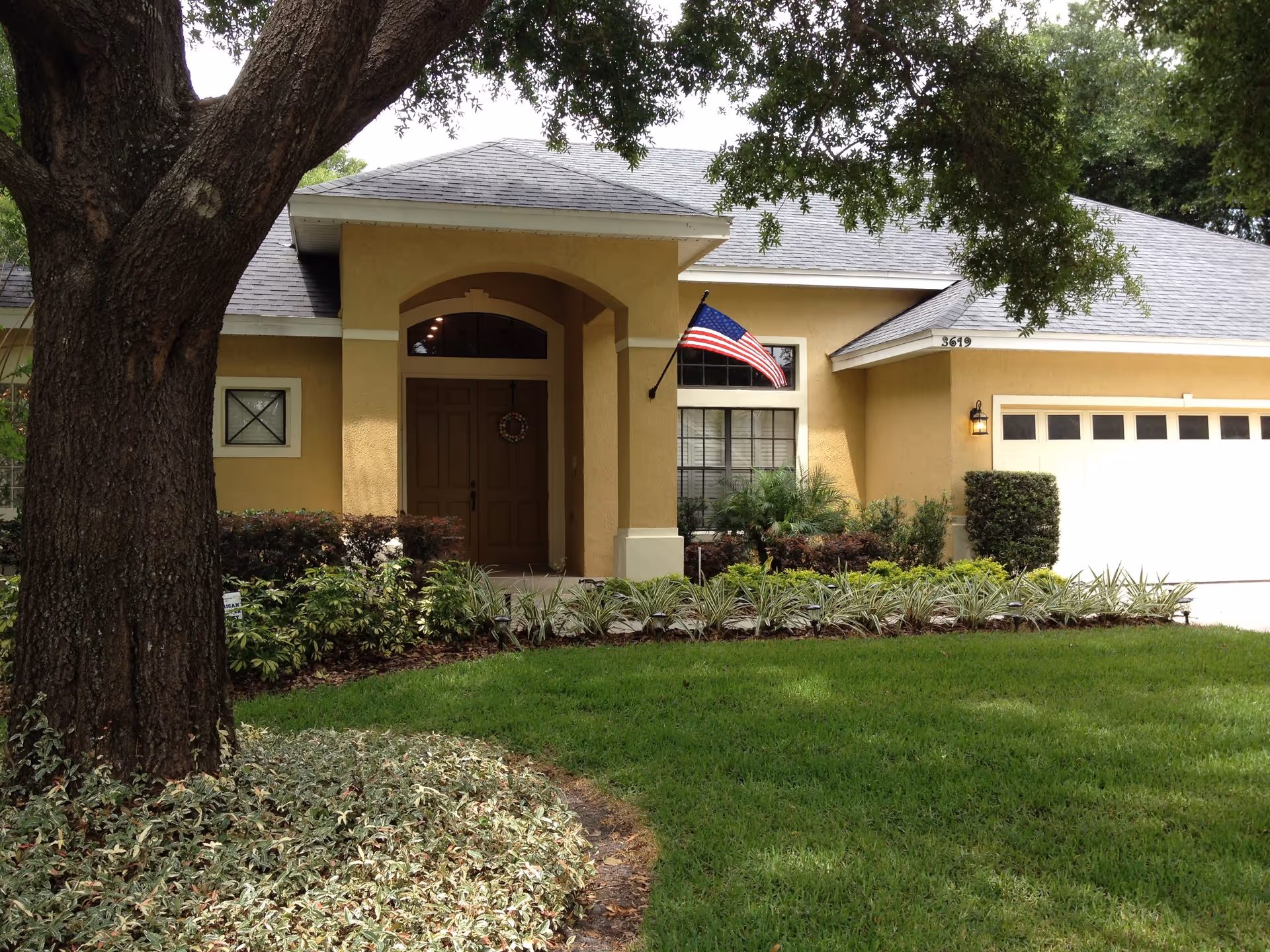 Front exterior of a single-story yellow house with a covered entry, American flag, garage, and landscaped lawn.