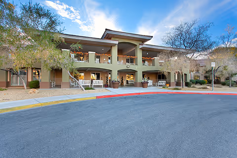 Two-story senior living building with covered balconies, an entrance canopy and a circular driveway under a blue sky.