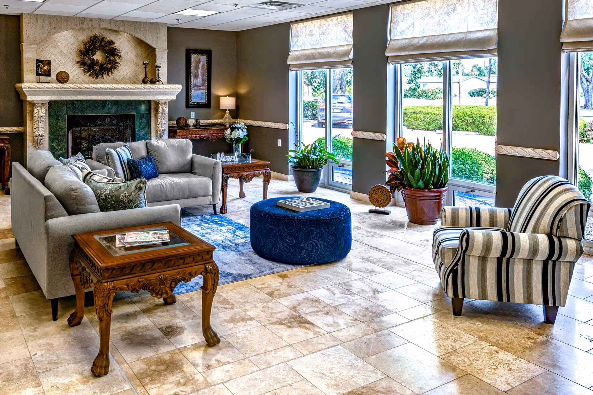 A cozy senior living facility common area with a gray sofa, a striped armchair, a blue round ottoman, and wooden side tables. The room features a decorative fireplace with a wreath above it, large windows with beige Roman shades, and several potted plants near the windows. The floor is tiled with light-colored stone.