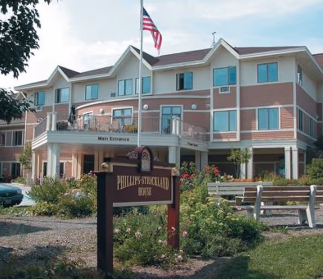 Front exterior of a three-story senior living building with a sign reading "Phillips Strickland House" and an American flag.