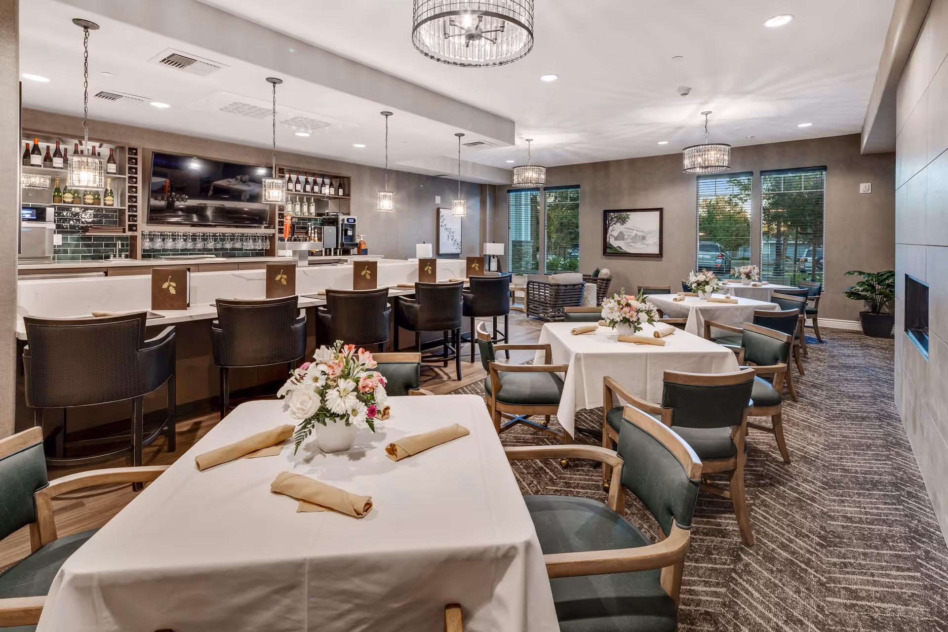 Well-lit dining area with multiple tables set with white tablecloths and floral centerpieces and a bar with high stools in the background.