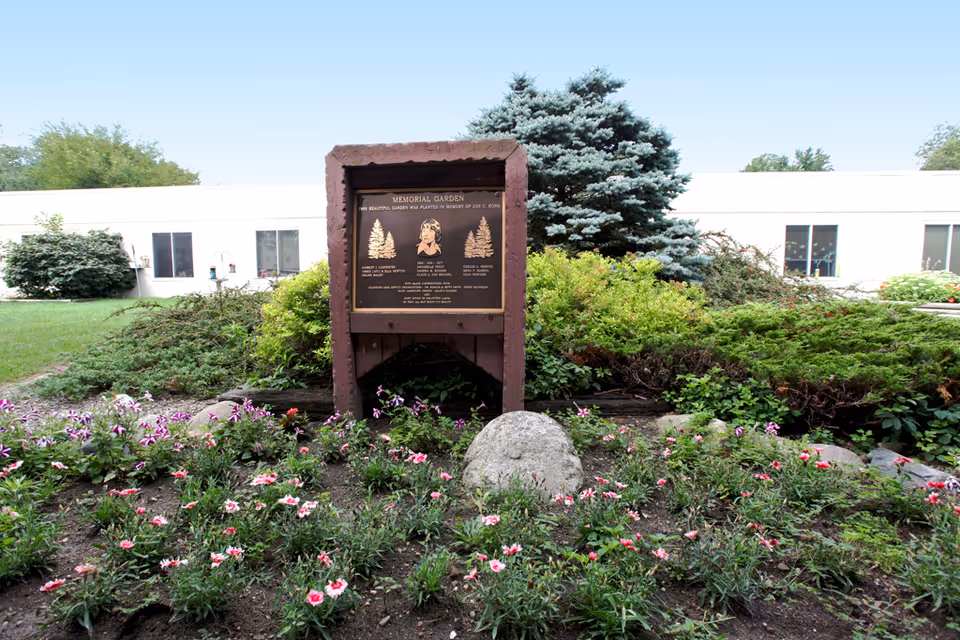 A memorial garden with a wooden plaque and flower beds in front of a low white building.