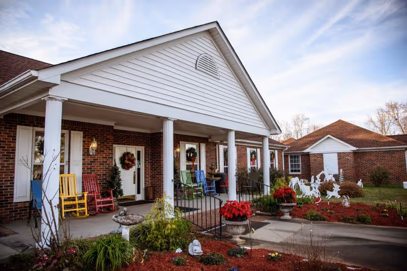 Covered front porch of a brick building with white columns, colorful rocking chairs, wreaths, and seasonal planters.