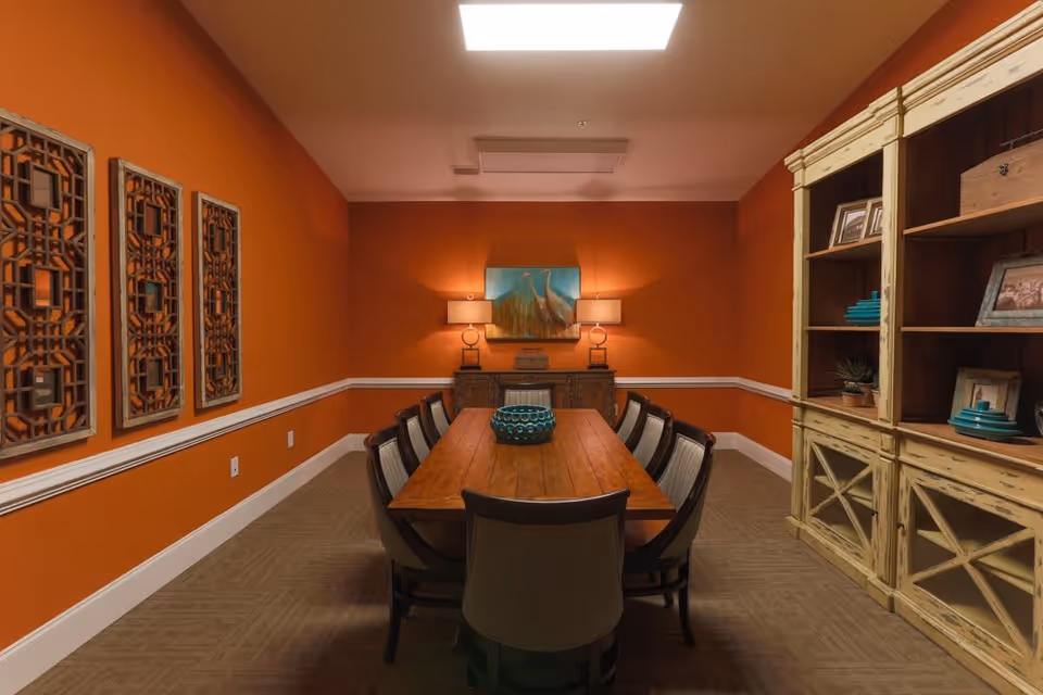 A dining room with orange walls and a long wooden table surrounded by eight chairs. On the table is a decorative blue bowl. The room features a wooden cabinet with shelves holding framed photos and decorative items on the right side. On the back wall, there is a sideboard with two lamps and a painting of birds above it. Three decorative wall panels are mounted on the left wall.