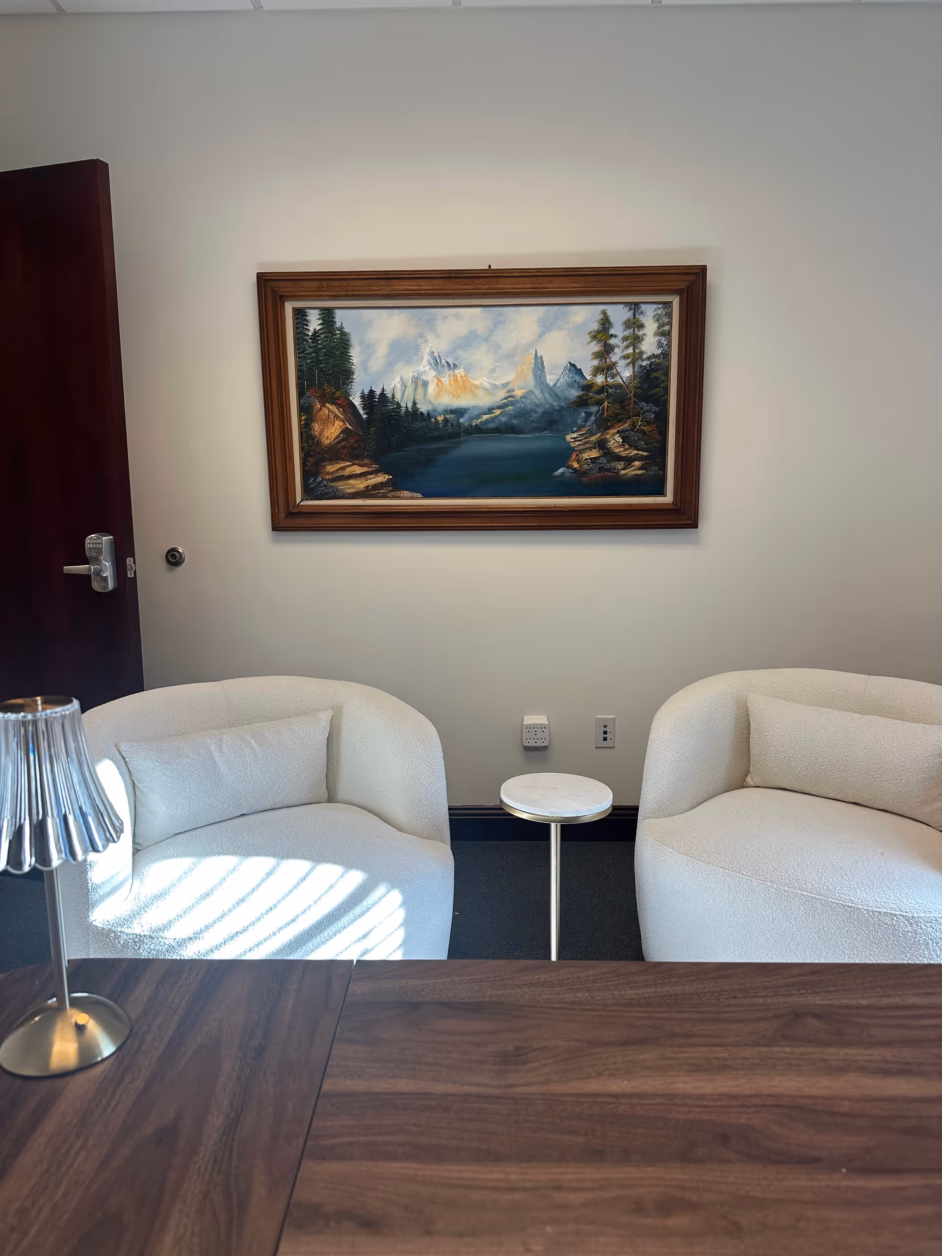 Two white curved chairs flank a small round table in a sunlit waiting area.
