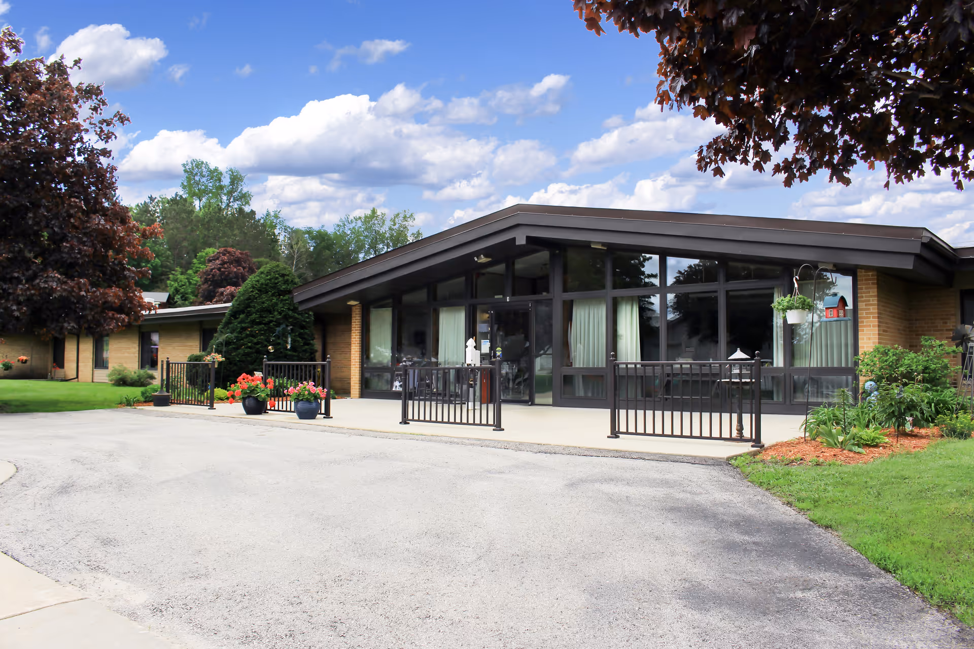 Exterior view of a single-story senior living facility building with large windows and a slanted roof. The entrance area is surrounded by black metal railings and decorated with potted flowers. Trees and green grass surround the building under a partly cloudy blue sky.