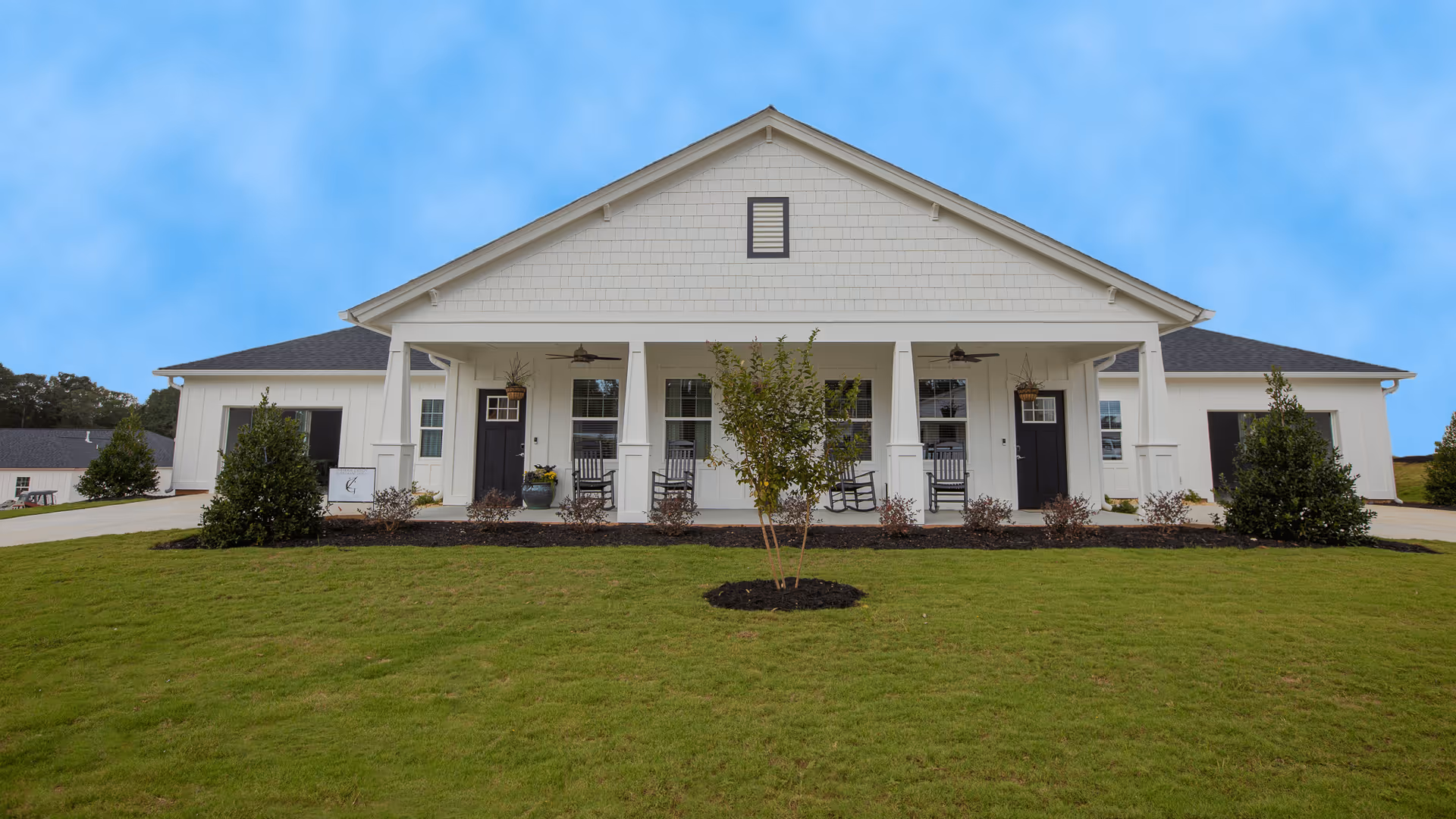 Front exterior view of a single-story building with white siding and a dark roof, featuring a covered porch with rocking chairs and ceiling fans. The building is surrounded by a well-maintained lawn with small trees and shrubs.