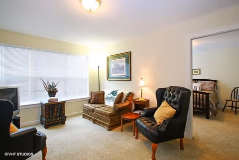 A cozy living room with a patterned sofa, a black leather armchair with a yellow pillow, a wooden side table with a lamp, and a floor lamp near a large window with blinds. A framed picture hangs on the wall above the sofa. The room has beige carpet and walls. An adjoining bedroom with a bed and a chair is visible through an open doorway.