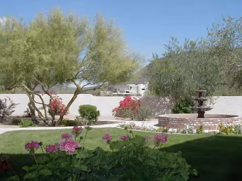 Sunlit outdoor courtyard with greenery, blooming flowers, and a brick fountain.