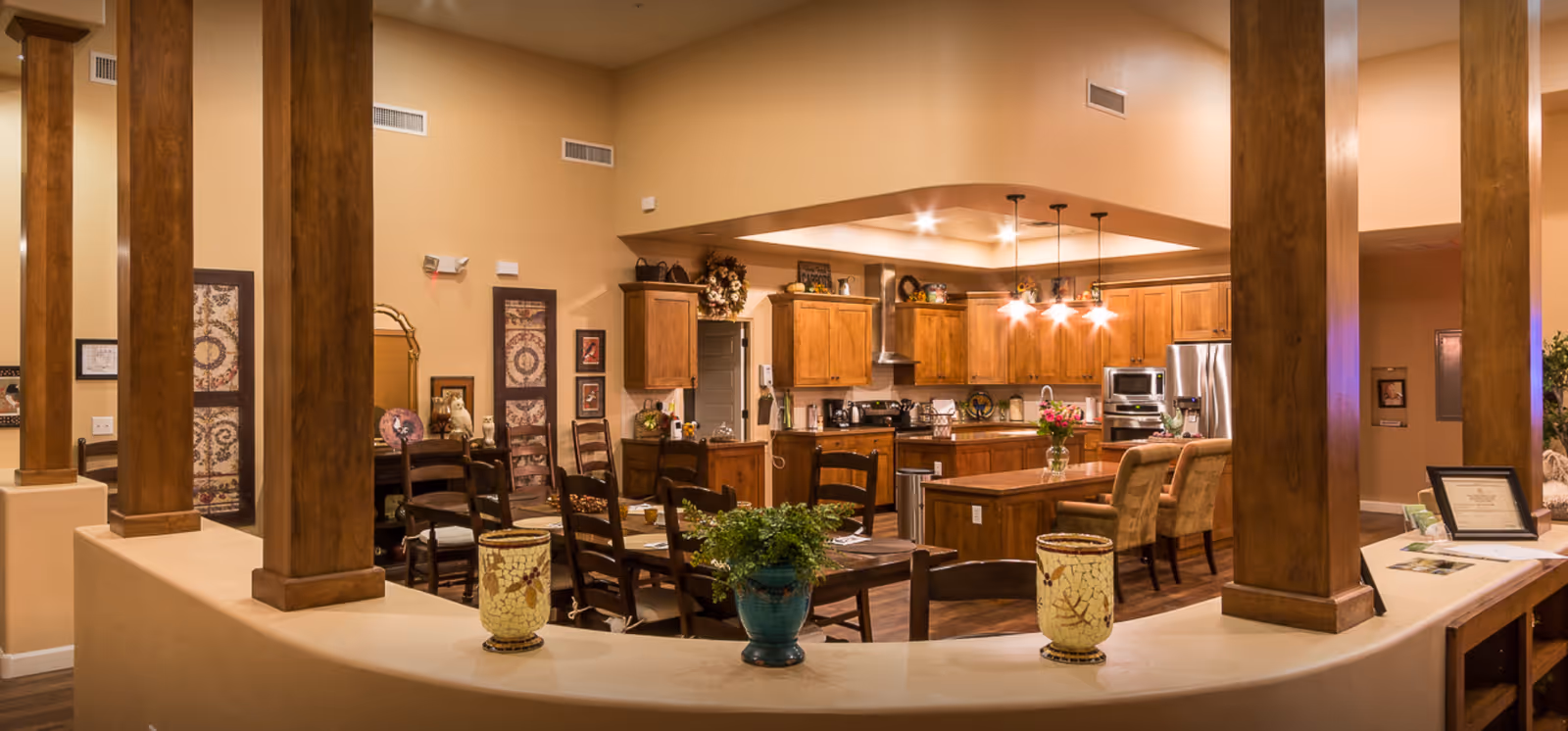 Senior living facility common area featuring a kitchen, dining tables, and wood-columned open layout.