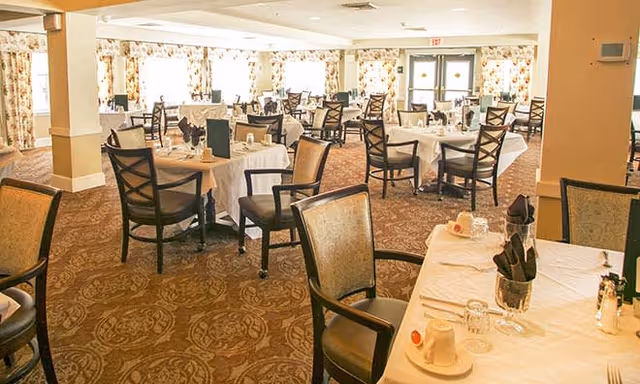 A spacious dining room with multiple tables covered in white tablecloths, each set with glasses, napkins, and menus. The room has patterned carpet flooring, large windows with floral curtains, and double doors at the far end.