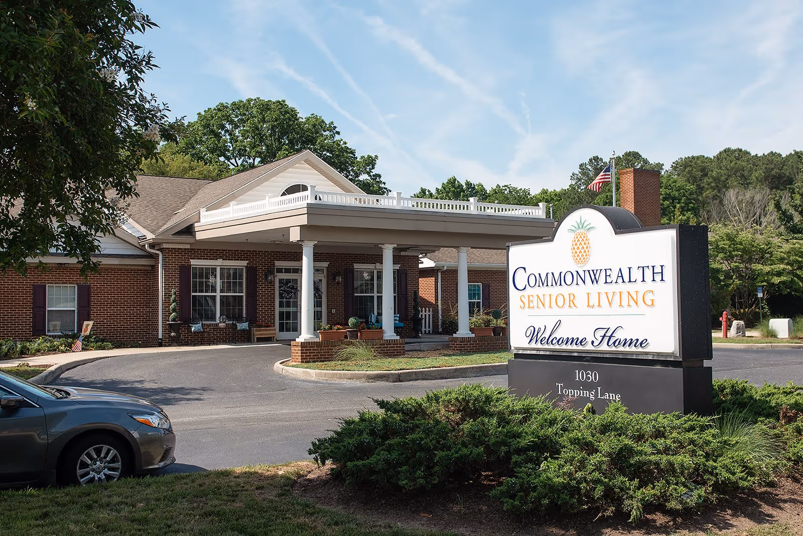 Exterior view of Commonwealth Senior Living at Hampton showing the entrance with a covered driveway, brick building, and a large sign with the facility name and address. There are trees and shrubs around the area and a car parked nearby.
