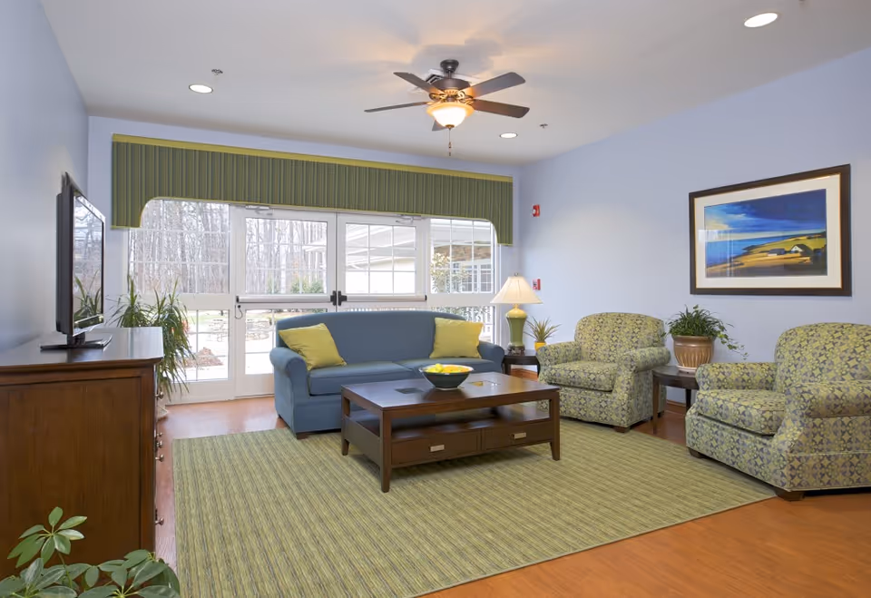 Bright senior living lounge with blue sofa, patterned chairs, coffee table, and TV.