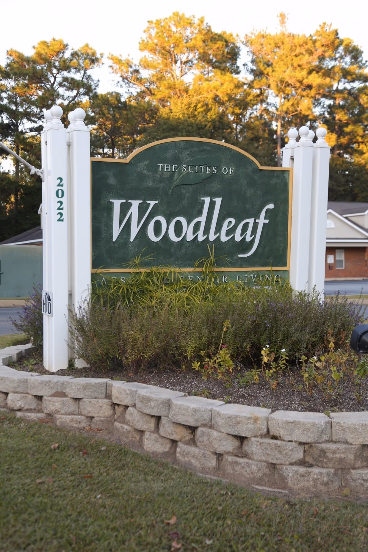 Entrance sign reading 'Woodleaf' surrounded by landscaping and stone edging in front of a senior living facility.