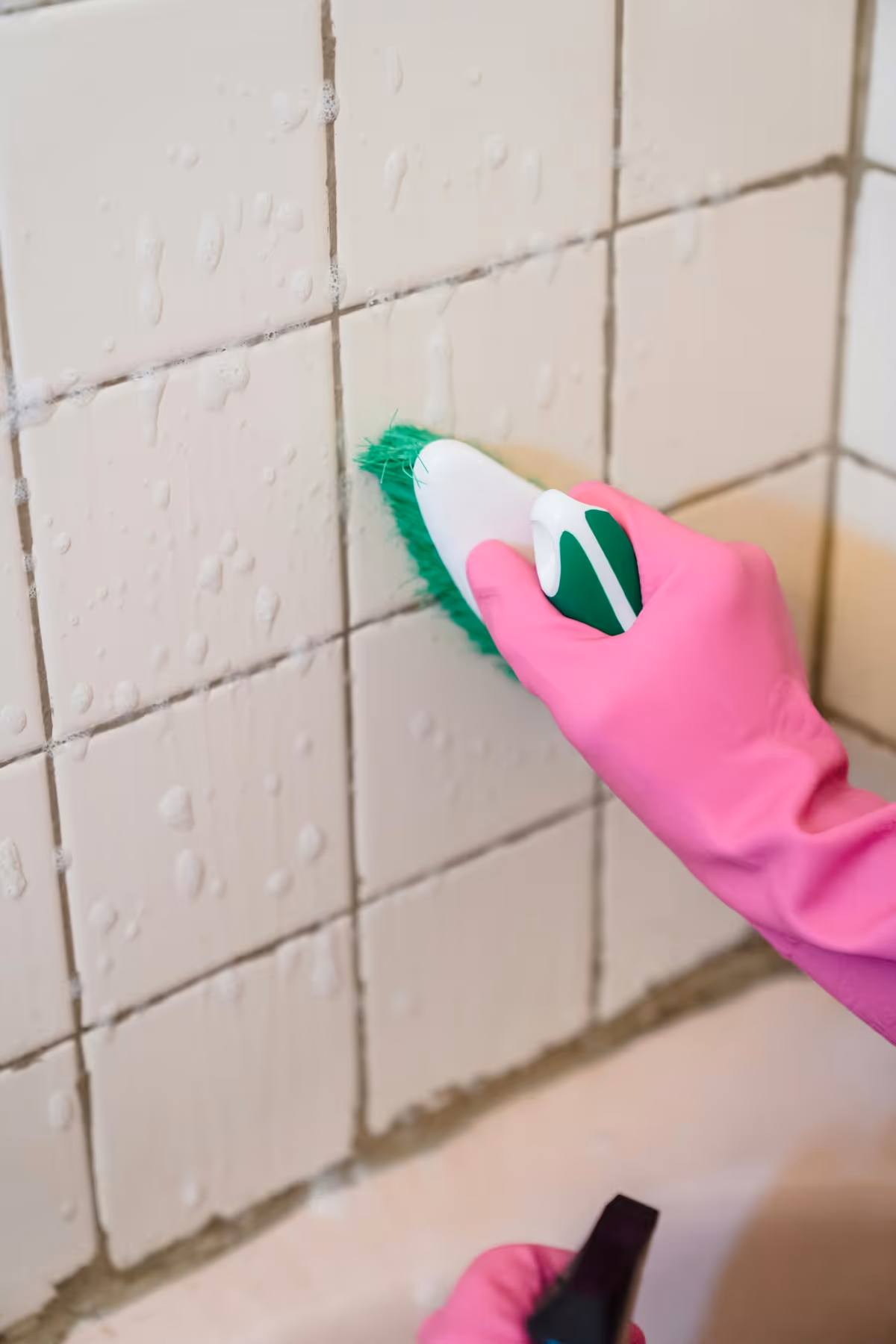 Gloved hand scrubs a tiled wall with a green scrubber and spray bottle.