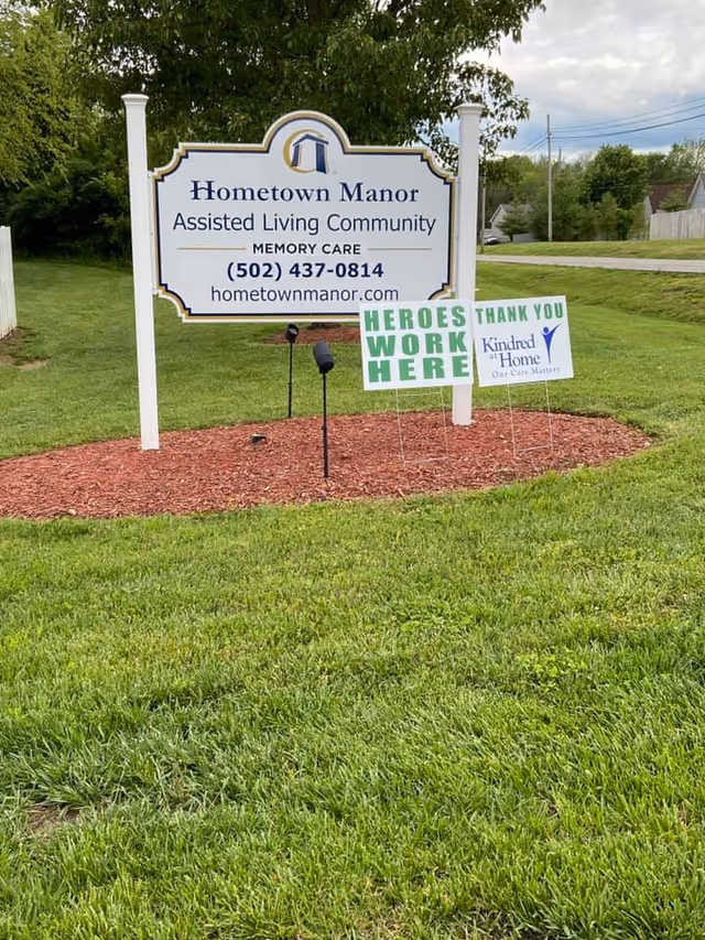 Outdoor view of a white sign for Hometown Manor Assisted Living Community with contact information and website, placed on a mulch bed surrounded by green grass. Two smaller signs in front read 'HEROES WORK HERE' and 'THANK YOU Kindred at Home Our Care Matters.' Trees and a road are visible in the background.