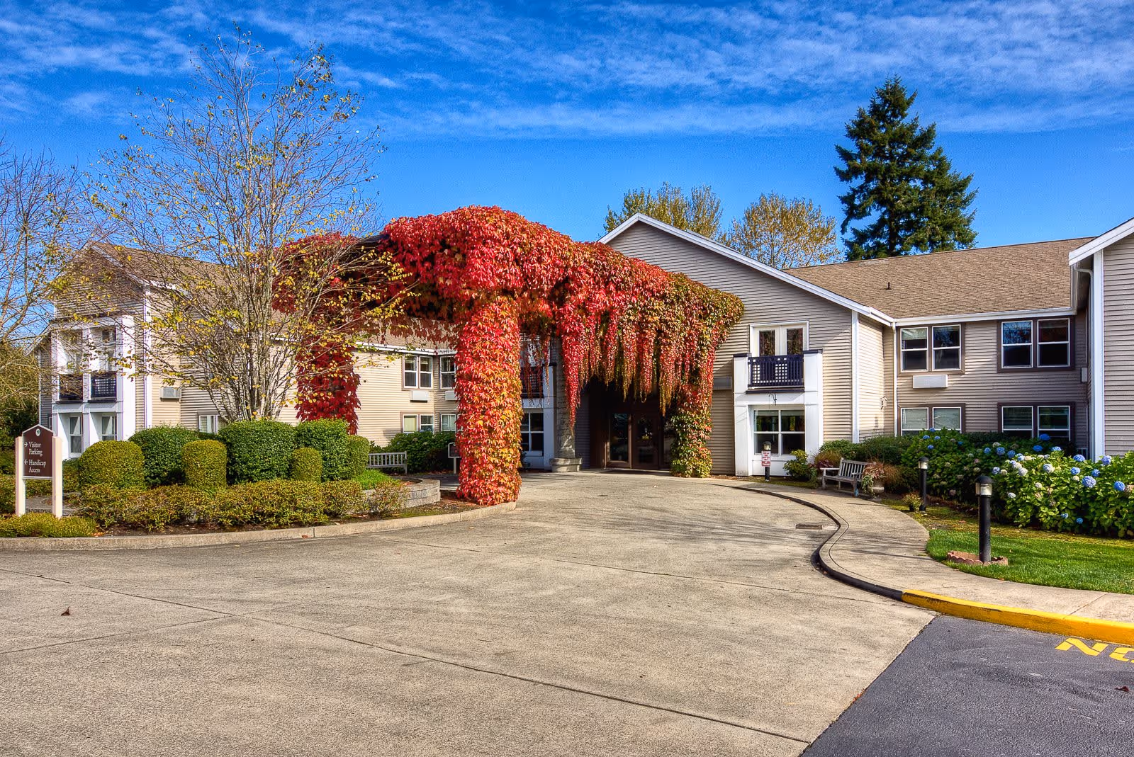 Exterior view of a senior living facility with a driveway leading to the entrance. The entrance is covered with vibrant red and green ivy hanging down from a pergola structure. The building is two stories with beige siding and several windows. There are well-maintained bushes, trees, and flower beds around the driveway under a clear blue sky.