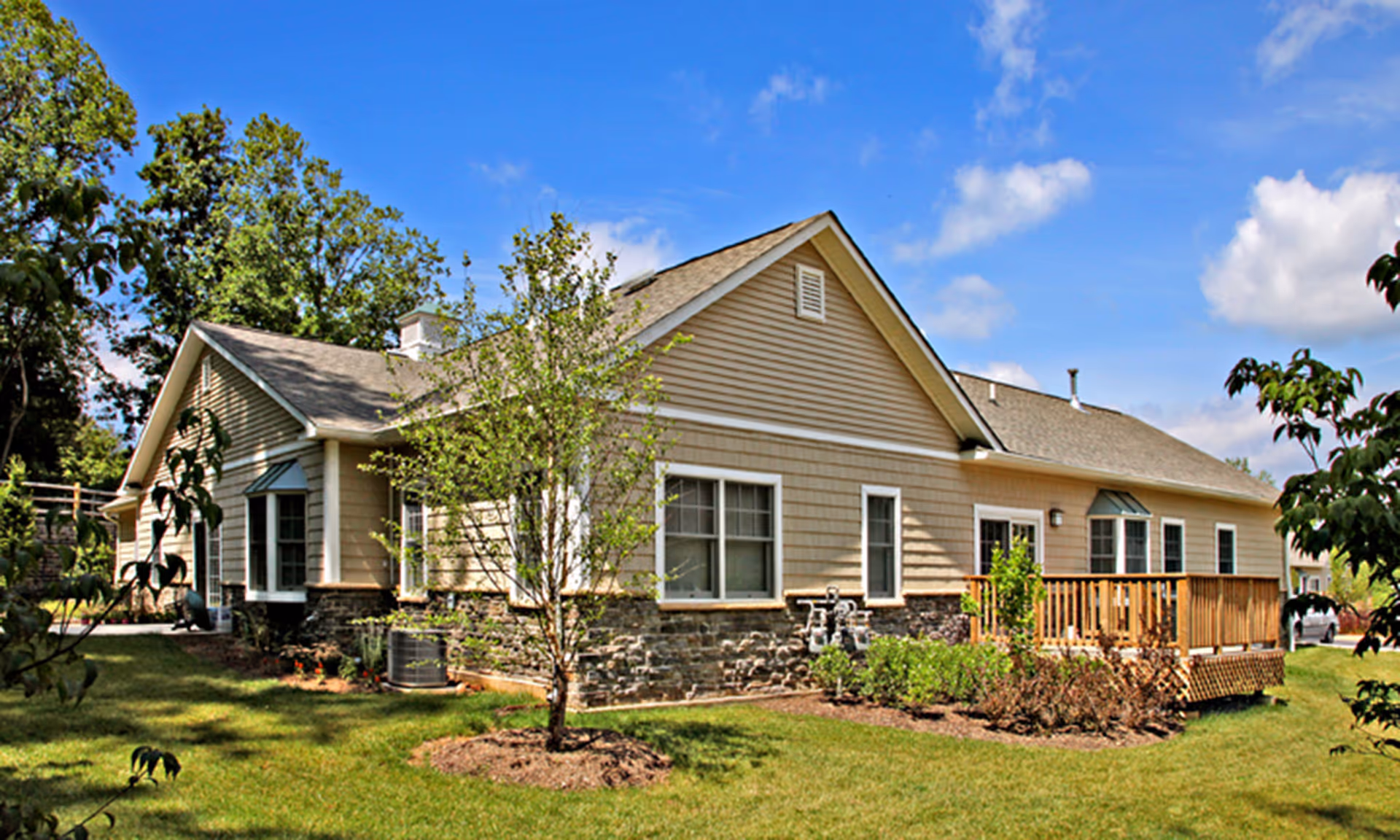 Single-story beige building with a small wooden deck, lawn, and surrounding trees under a blue sky.