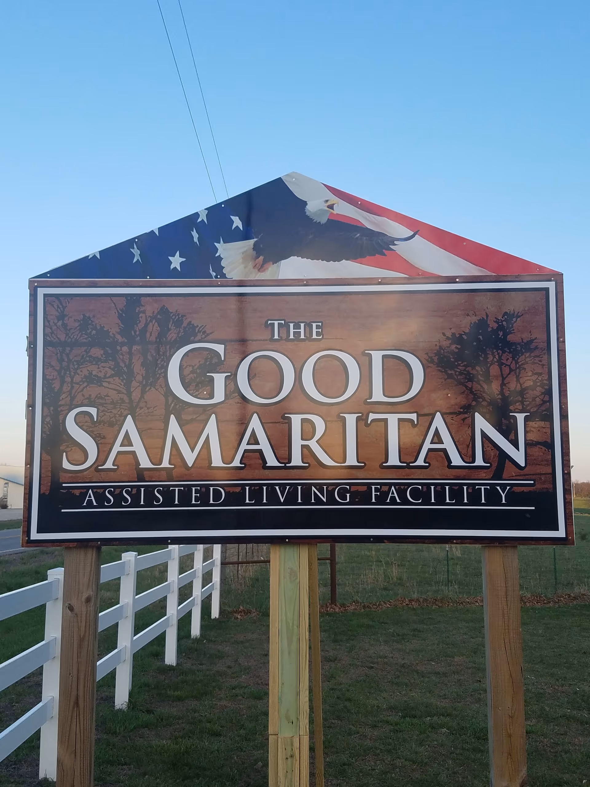 A large wooden sign for The Good Samaritan Assisted Living Facility with an American flag and bald eagle design at the top, set outdoors with a white fence and grassy area in the background under a clear blue sky.