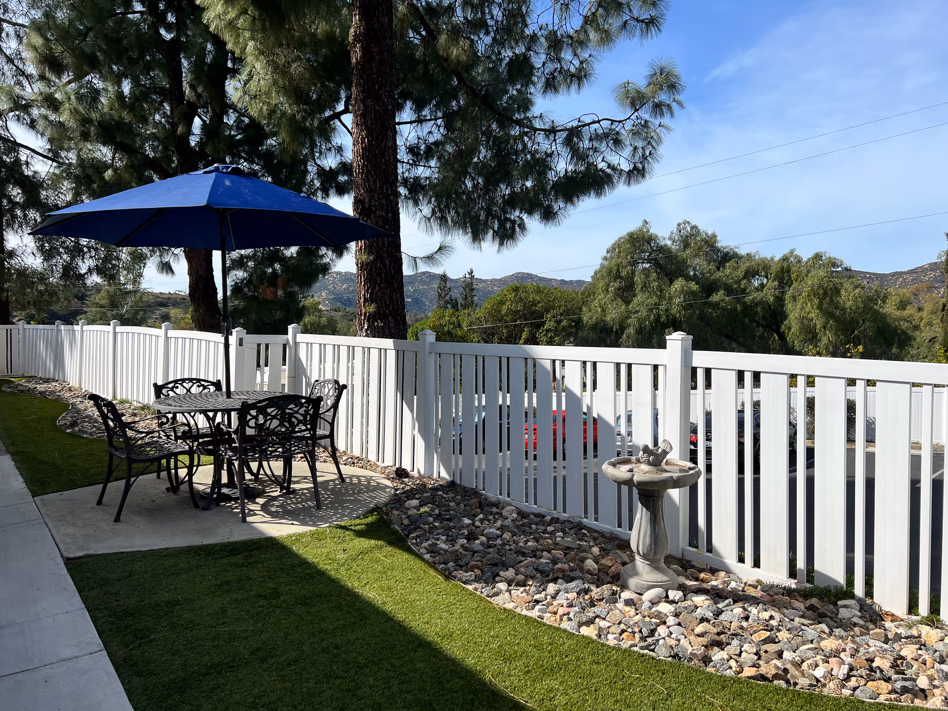 Outdoor seating area with a black metal table and four chairs under a blue umbrella, next to a white fence. There is a small birdbath on a bed of rocks beside the fence, with trees and hills visible in the background under a clear blue sky.