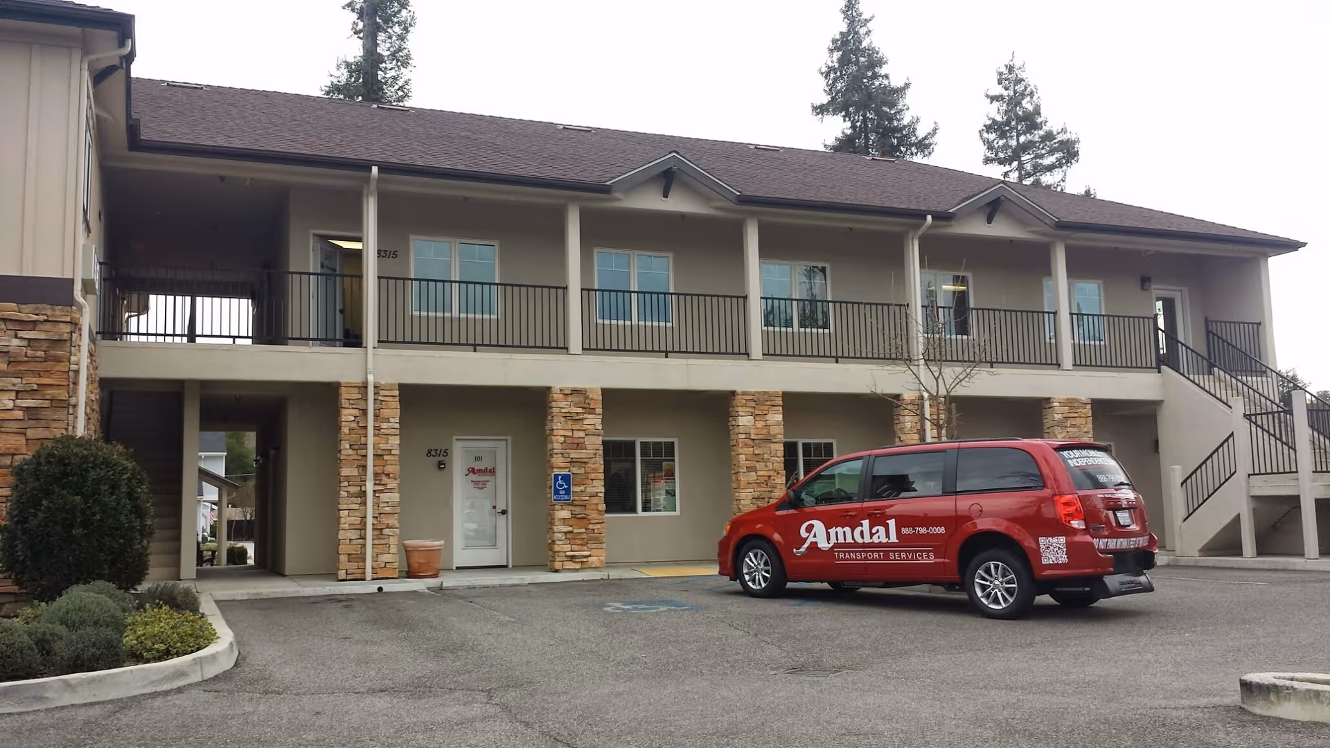 Two-story beige building with stone pillars, railing balcony, and a red van parked front.