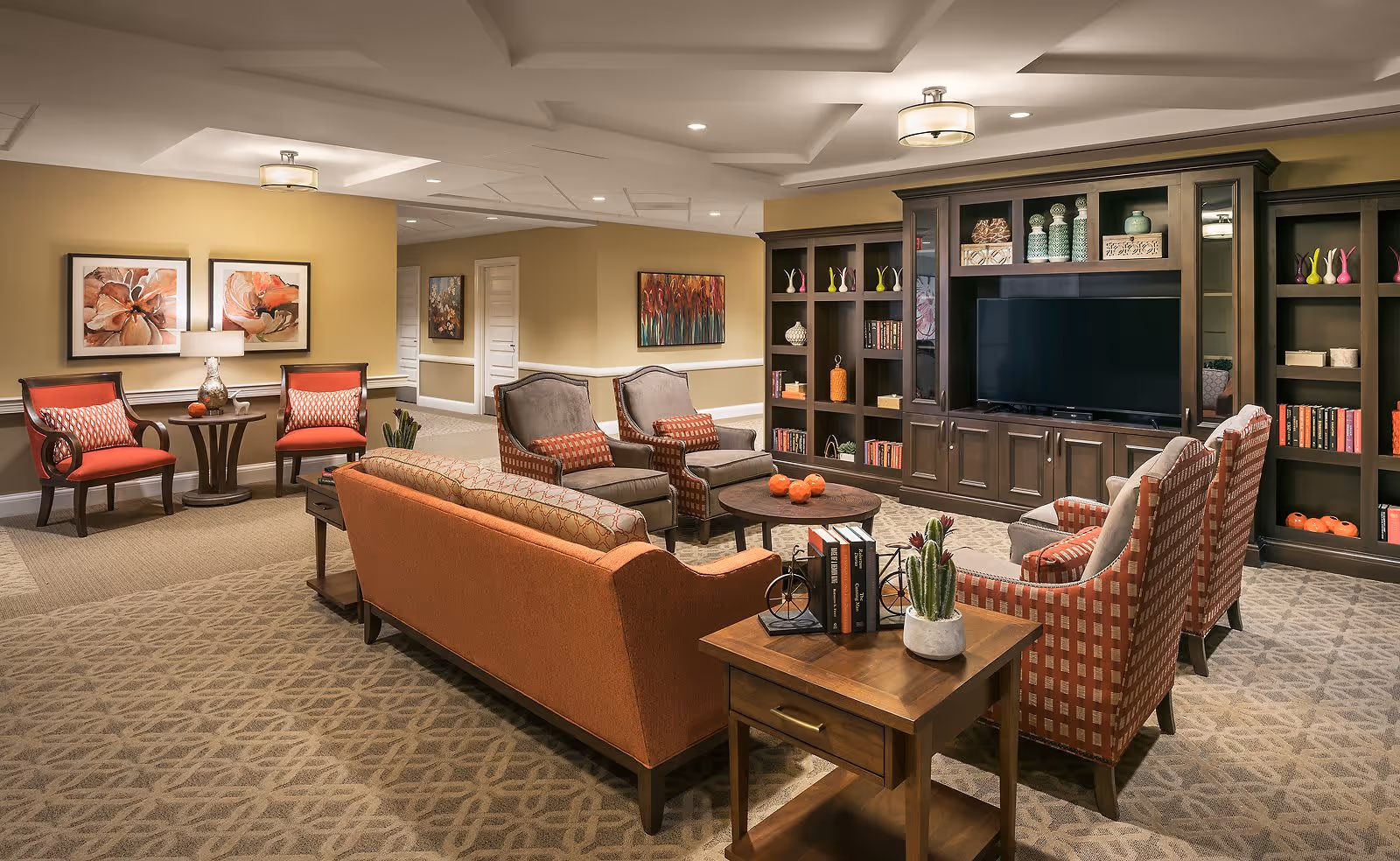 A cozy and well-decorated living room area in a senior living facility featuring an orange sofa, several upholstered armchairs with patterned cushions, a wooden coffee table with decorative orange spheres, and a large dark wood entertainment center with a flat-screen TV and shelves filled with books and decorative items. The room has beige walls adorned with framed artwork and soft lighting from ceiling fixtures.