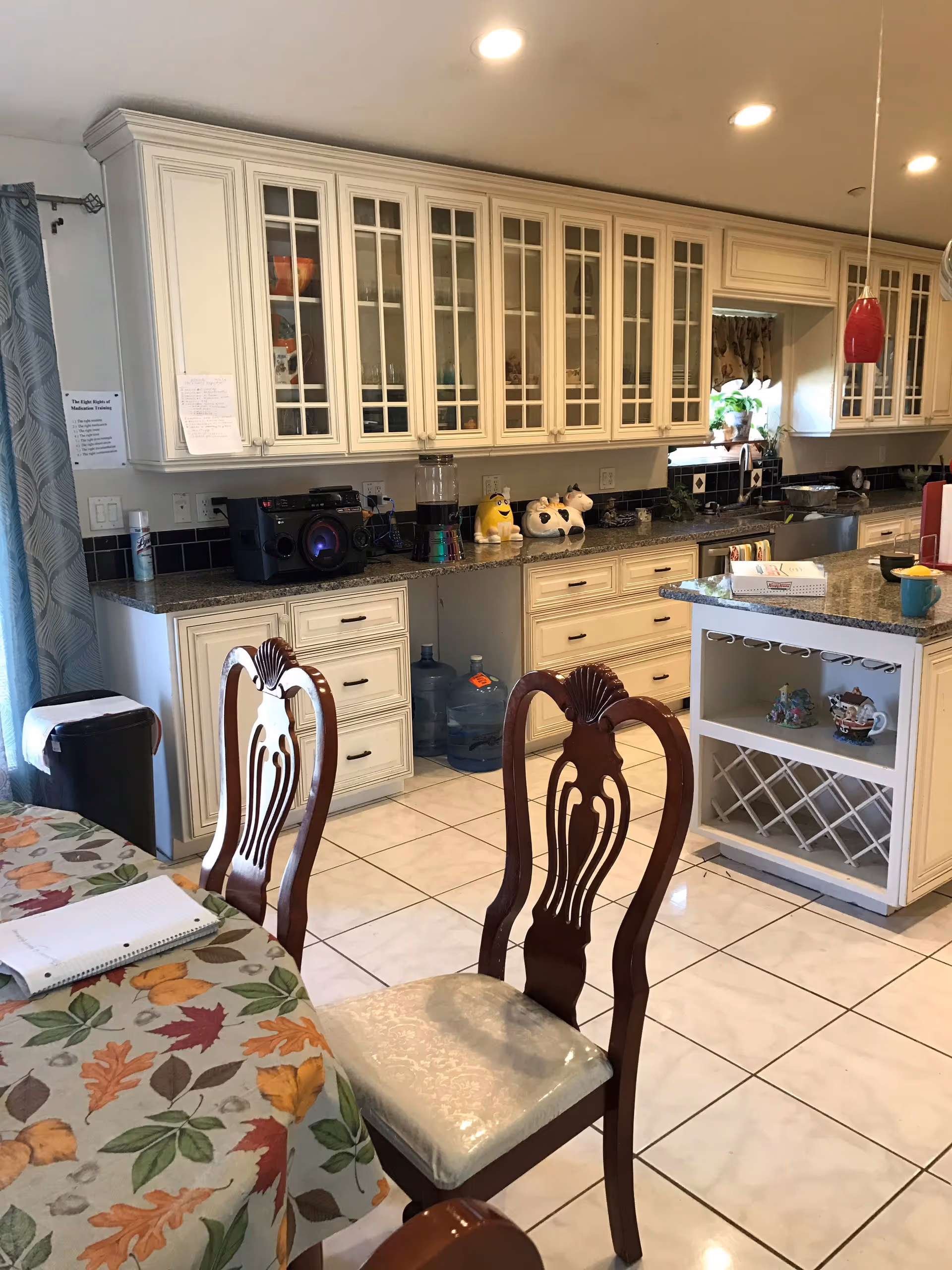 A spacious kitchen with cream-colored cabinets featuring glass doors, a granite countertop, and a kitchen island with open shelving and a wine rack. Two wooden chairs with cushioned seats are positioned near a table covered with a floral tablecloth. Various items including a notebook, a radio, decorative containers, and water jugs are visible on the countertops. The floor is tiled in white, and recessed ceiling lights illuminate the room.