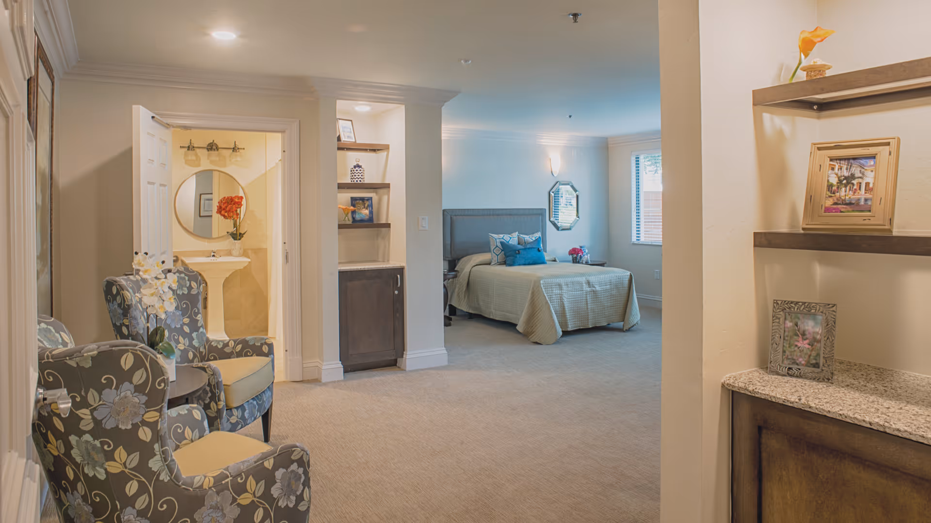A spacious senior living bedroom with a bed, bedside table, and window with blinds. To the left, there is an open door leading to a bathroom with a pedestal sink and round mirror. Two floral-patterned armchairs with a small table between them are positioned near the bathroom door. Shelves with framed pictures and decorative items are visible on the right side of the image.
