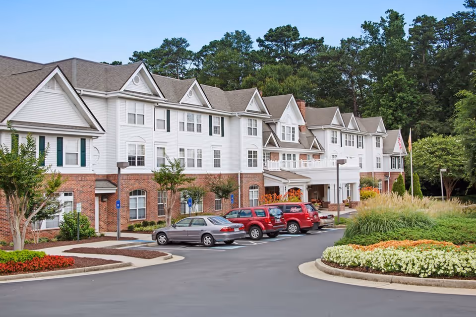 Exterior view of a large senior living facility building with white siding and brick accents. Several cars are parked in front of the building, which is surrounded by landscaped greenery and flower beds. Trees are visible in the background under a clear sky.