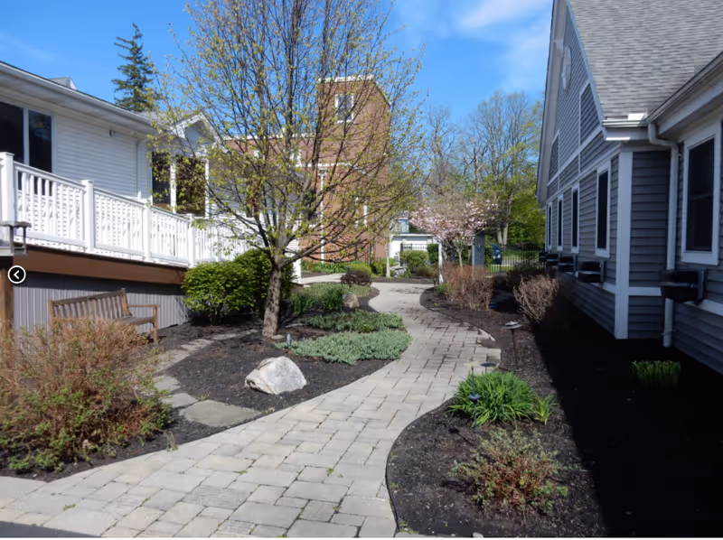 A winding stone walkway through a landscaped courtyard with a bench between two senior living buildings.