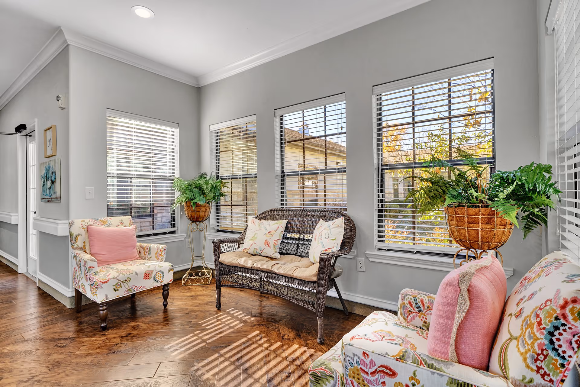 A bright and cozy sitting area with three windows letting in natural light. The room features two floral patterned armchairs with pink cushions, a wicker loveseat with beige cushions and floral pillows, and two potted plants on stands near the windows. The walls are painted light gray and the floor is wooden.