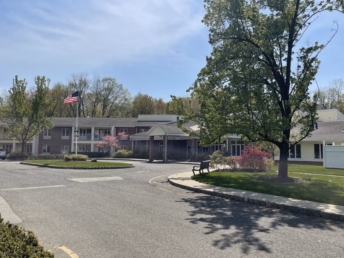 Exterior view of Mira Vie at Montville assisted living facility showing a two-story building with balconies, an American flag on a flagpole, a large tree, a bench, and landscaped greenery under a partly cloudy sky.