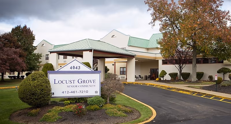 Exterior view of Locust Grove Senior Community building with a covered entrance, surrounded by landscaped bushes and trees with autumn foliage under a cloudy sky.