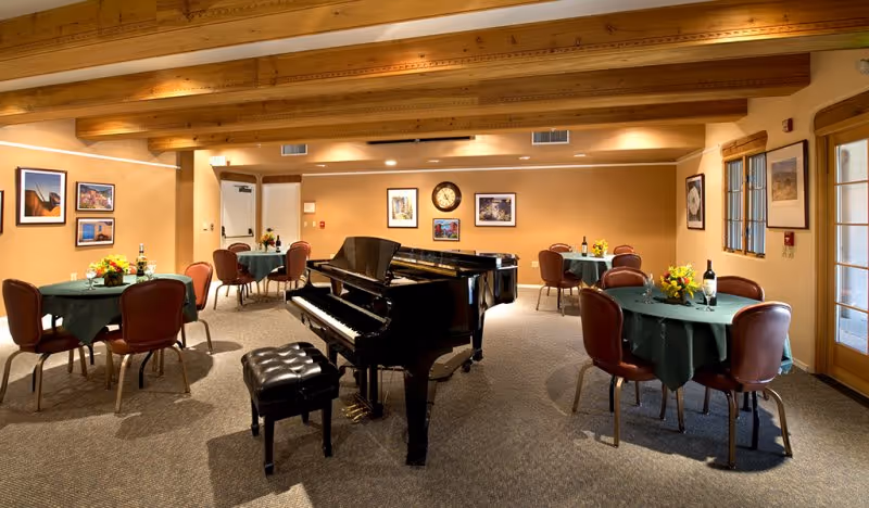 Interior room with a black grand piano in the center surrounded by round tables covered with green tablecloths and brown chairs. The tables have flower arrangements and wine bottles. The room has wooden ceiling beams, framed pictures on the walls, a clock, and large windows on one side.