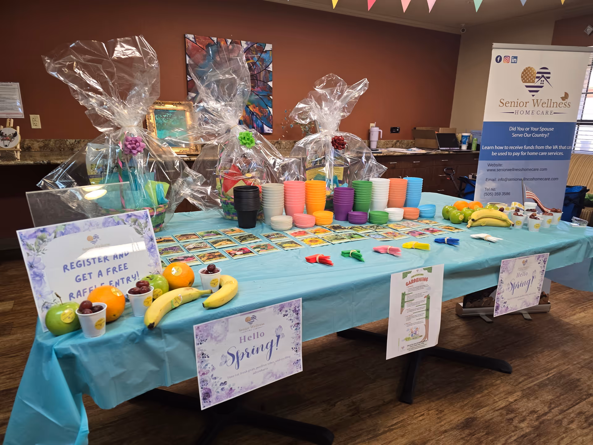 Colorful event table with wrapped gift baskets, bowls, and a Senior Wellness Home Care banner indoors.
