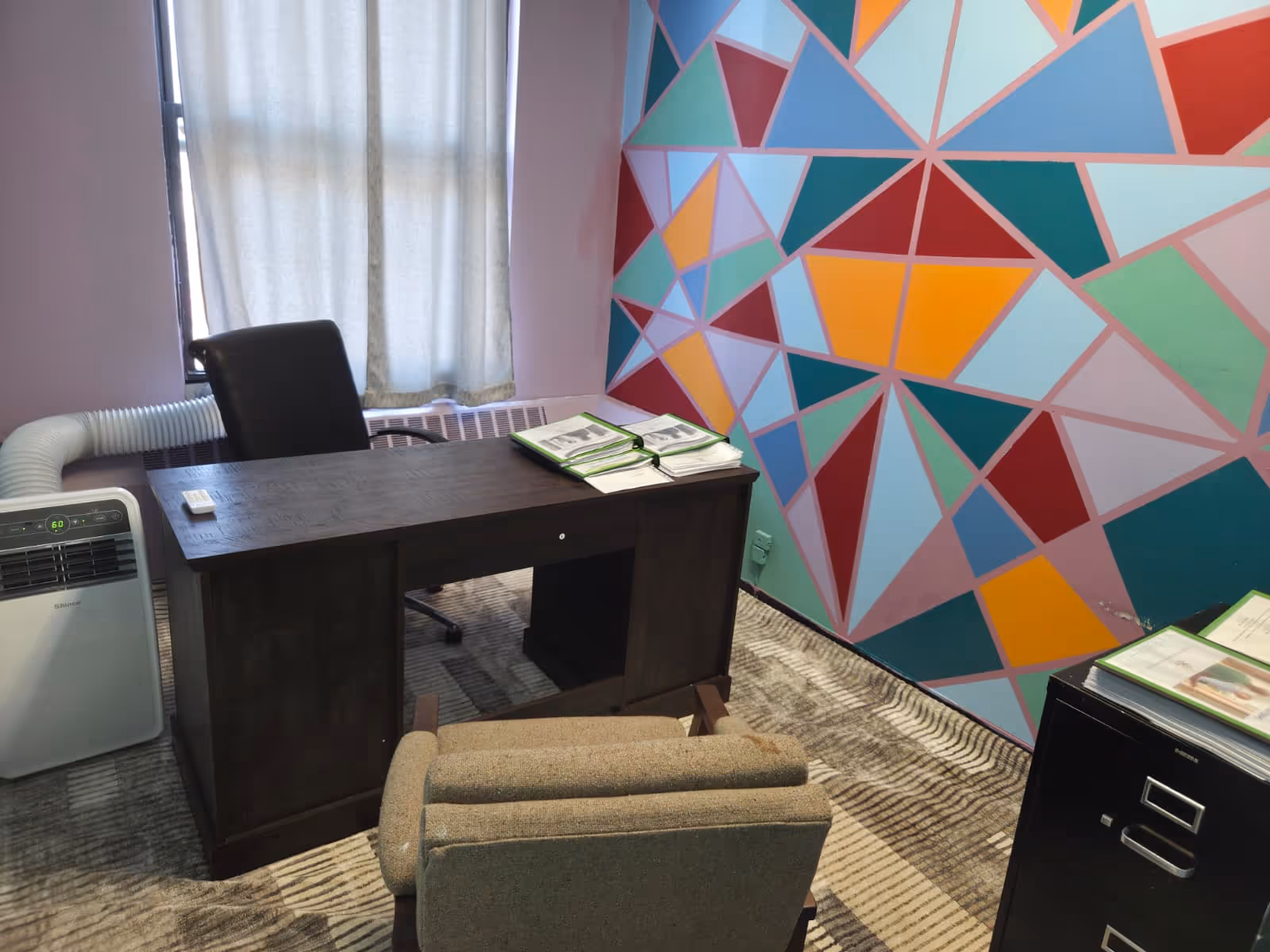 A small office with a dark desk, colorful geometric wall, and filing cabinet.