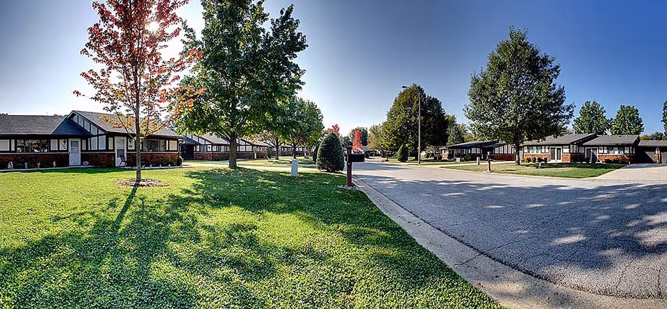 Panoramic exterior view of one-story residential villas with green lawns, trees, and a paved road in a senior living community.