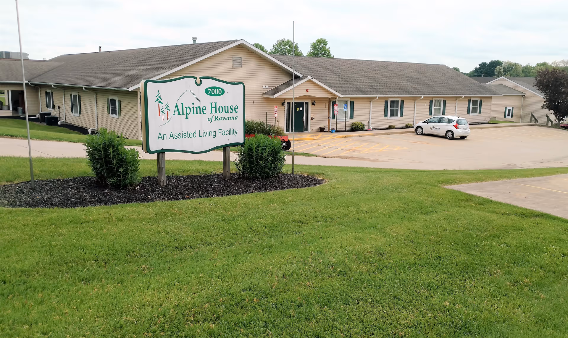 Exterior front view of Alpine House of Ravenna assisted living facility showing the entrance, parking lot, and a large sign on the lawn.